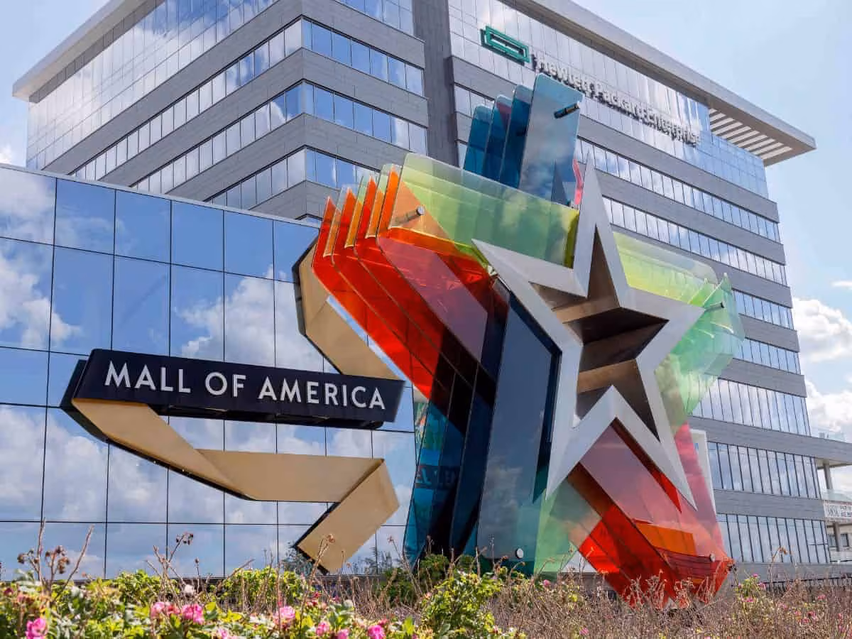 The main entrance to Mall of America features the iconic multicolored star sculpture in front of the building. A black Mall of America sign extends from the left side of the sculpture. The glass-front building reflects the blue sky and clouds, with flowers and landscaping visible in the foreground.