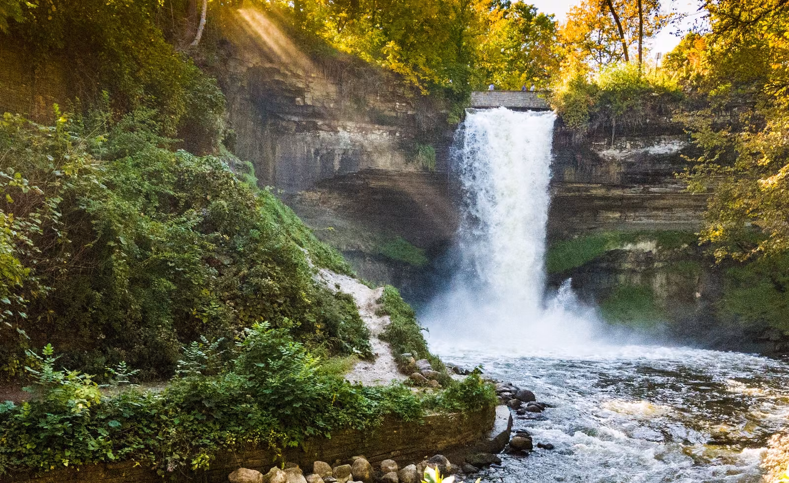 Minnehaha Falls cascades over a limestone cliff into the river below, surrounded by lush green trees and vegetation. Sunlight filters through the trees, creating rays of light across the waterfall. A stone bridge crosses the top of the falls, with a few visitors enjoying the view.