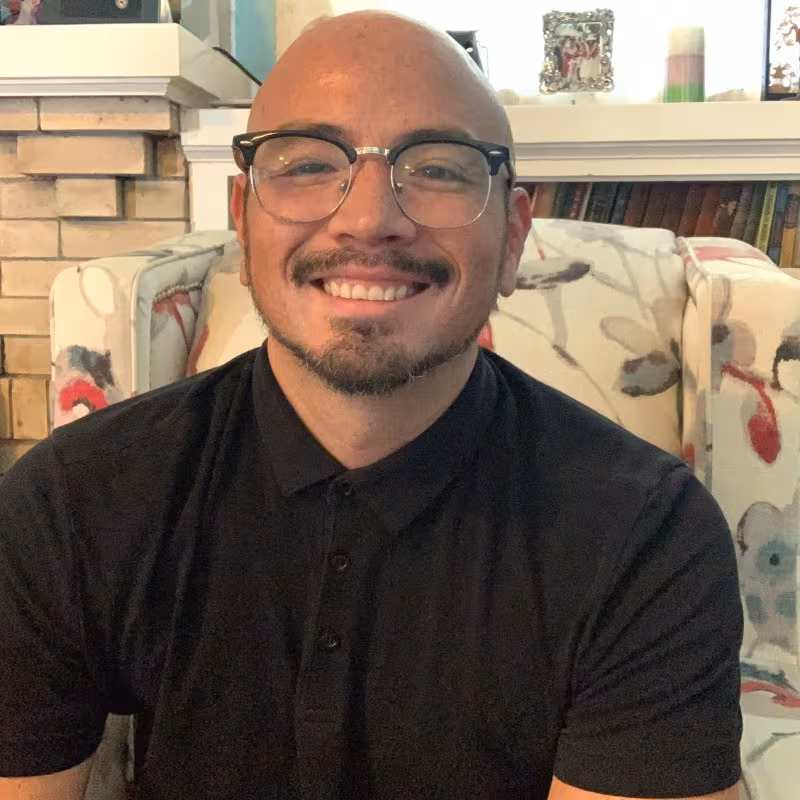 Bald man with glasses and neatly groomed facial hair, smiling in a black collared shirt in a cozy living room with a brick fireplace and patterned chair behind him.